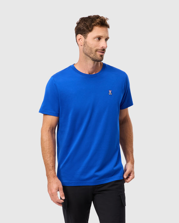 A man with short hair and a beard is wearing a Psycho Bunny MENS CLASSIC CREW NECK TEE in bright blue, crafted from soft Pima cotton, featuring an embroidered logo on the chest. He is looking to the side while wearing black pants against a light gray background.