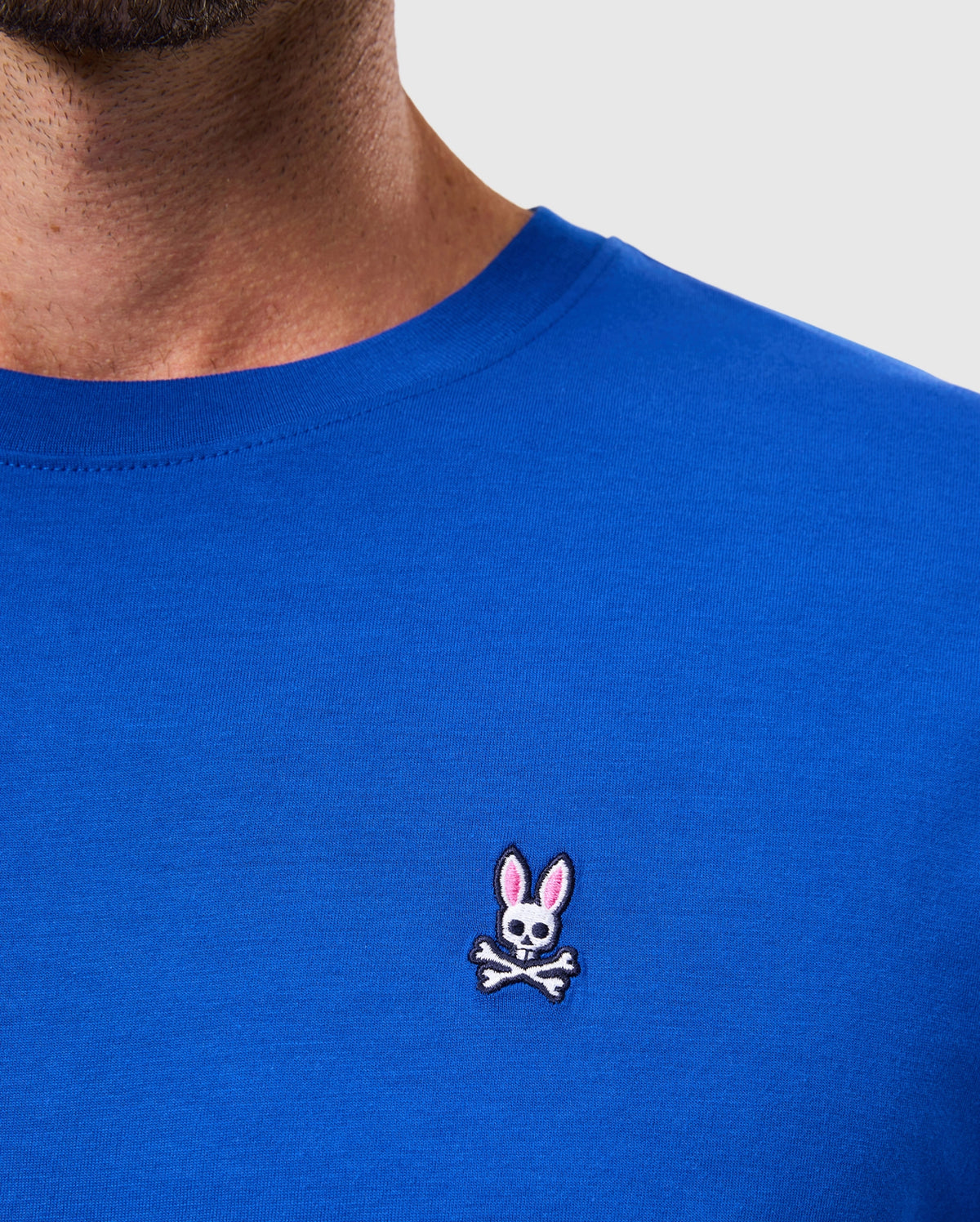 A person wearing the MENS CLASSIC CREW NECK TEE - B6U014CRPC by Psycho Bunny, crafted from soft Pima cotton in blue, featuring a small embroidered logo of a skull with bunny ears above crossed bones.