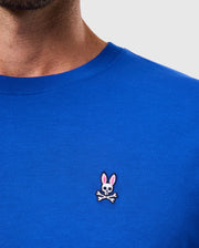 A person wearing the MENS CLASSIC CREW NECK TEE - B6U014CRPC by Psycho Bunny, crafted from soft Pima cotton in blue, featuring a small embroidered logo of a skull with bunny ears above crossed bones.