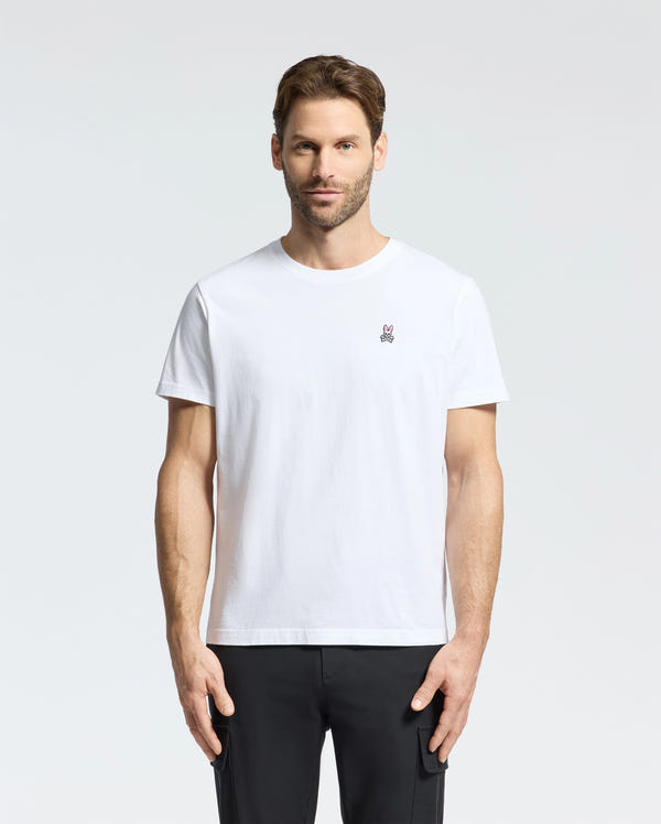 A man with short brown hair and a beard wears a Psycho Bunny MENS CLASSIC CREW NECK TEE - B6U014CRPC, crafted from soft Pima cotton, featuring a small embroidered logo. Paired with black pants, he stands against a light gray background, showcasing this men's wardrobe staple.