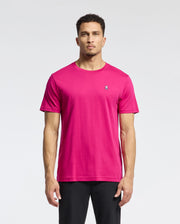 Against a white backdrop, a man wears the Psycho Bunny Men's Classic Crew Neck Tee in bright pink, made from premium Pima cotton. It features an embroidered bunny logo on the chest. His short curly hair complements this look, paired with sleek black pants.