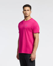 A man wears a Psycho Bunny bright pink MENS CLASSIC CREW NECK TEE (B6U014CRPC) featuring a small embroidered Bunny logo on the chest. Standing against a white background, he has short curly hair and looks directly at the camera, paired with sleek black pants.