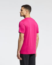 A person with short curly hair stands sideways in a bright pink Psycho Bunny MENS CLASSIC CREW NECK TEE (B6U014CRPC) made of Pima cotton and black pants, with an embroidered Bunny logo on the shirt's front against a plain white background.