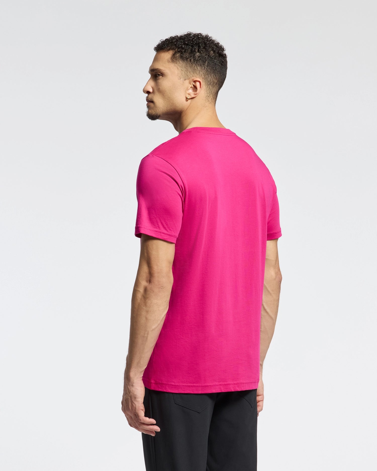 A person with short curly hair stands sideways in a bright pink Psycho Bunny MENS CLASSIC CREW NECK TEE (B6U014CRPC) made of Pima cotton and black pants, with an embroidered Bunny logo on the shirt's front against a plain white background.