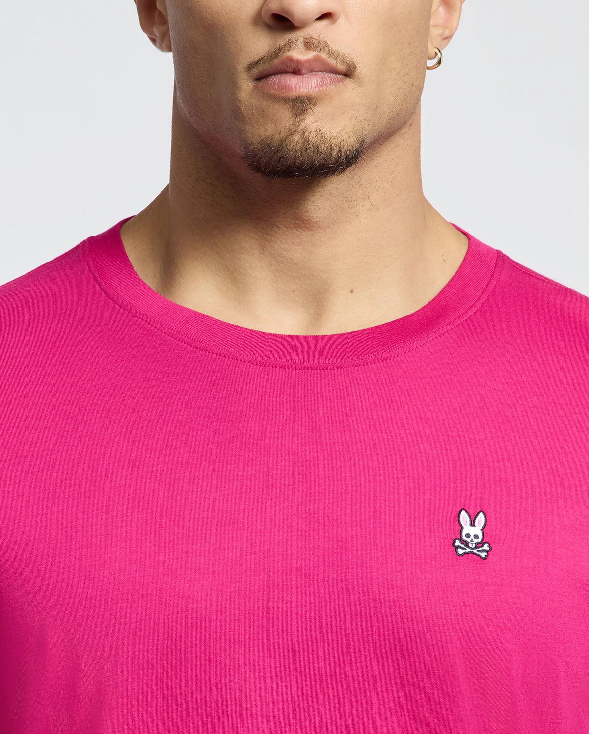 The individual is sporting a Psycho Bunny MENS CLASSIC CREW NECK TEE - B6U014CRPC in bright pink Pima cotton. The embroidered bunny logo with crossed bones on the chest complements their short beard and hoop earrings, all set against a plain white background.