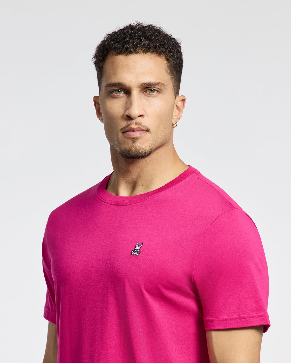 A man with short curly hair and an earring wears a bright pink Psycho Bunny MENS CLASSIC CREW NECK TEE - B6U014CRPC, crafted from soft Pima cotton boasting an embroidered bunny logo on the chest, against a plain light grey background.
