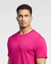 A man with short curly hair and an earring wears a bright pink Psycho Bunny MENS CLASSIC CREW NECK TEE - B6U014CRPC, crafted from soft Pima cotton boasting an embroidered bunny logo on the chest, against a plain light grey background.