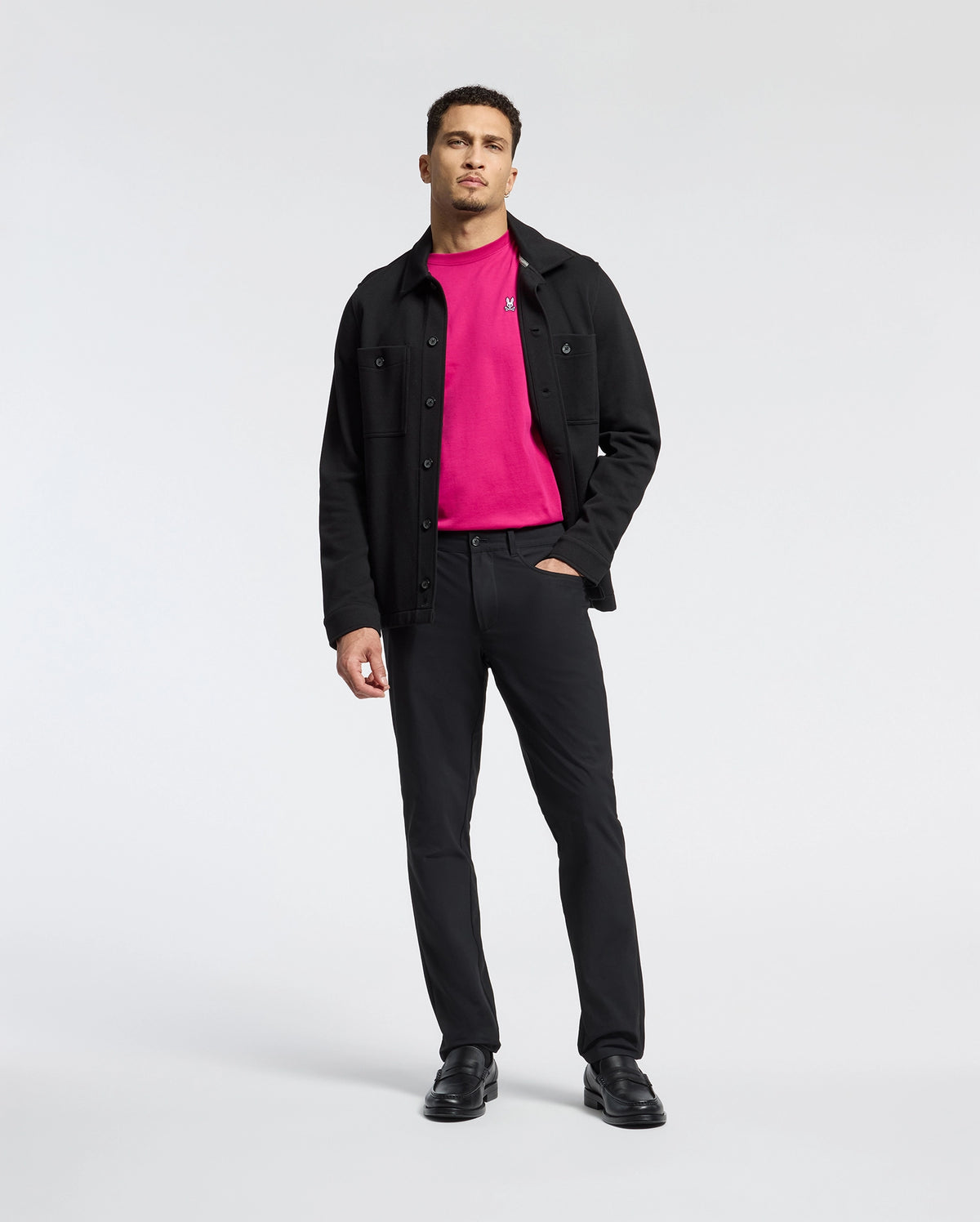 A man in a Psycho Bunny MENS CLASSIC CREW NECK TEE - B6U014CRPC, beneath a black jacket, with matching pants and shoes, stands against a white background. His left hand is in his pocket as he gazes directly at the camera.