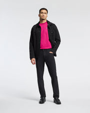 A man in a Psycho Bunny MENS CLASSIC CREW NECK TEE - B6U014CRPC, beneath a black jacket, with matching pants and shoes, stands against a white background. His left hand is in his pocket as he gazes directly at the camera.