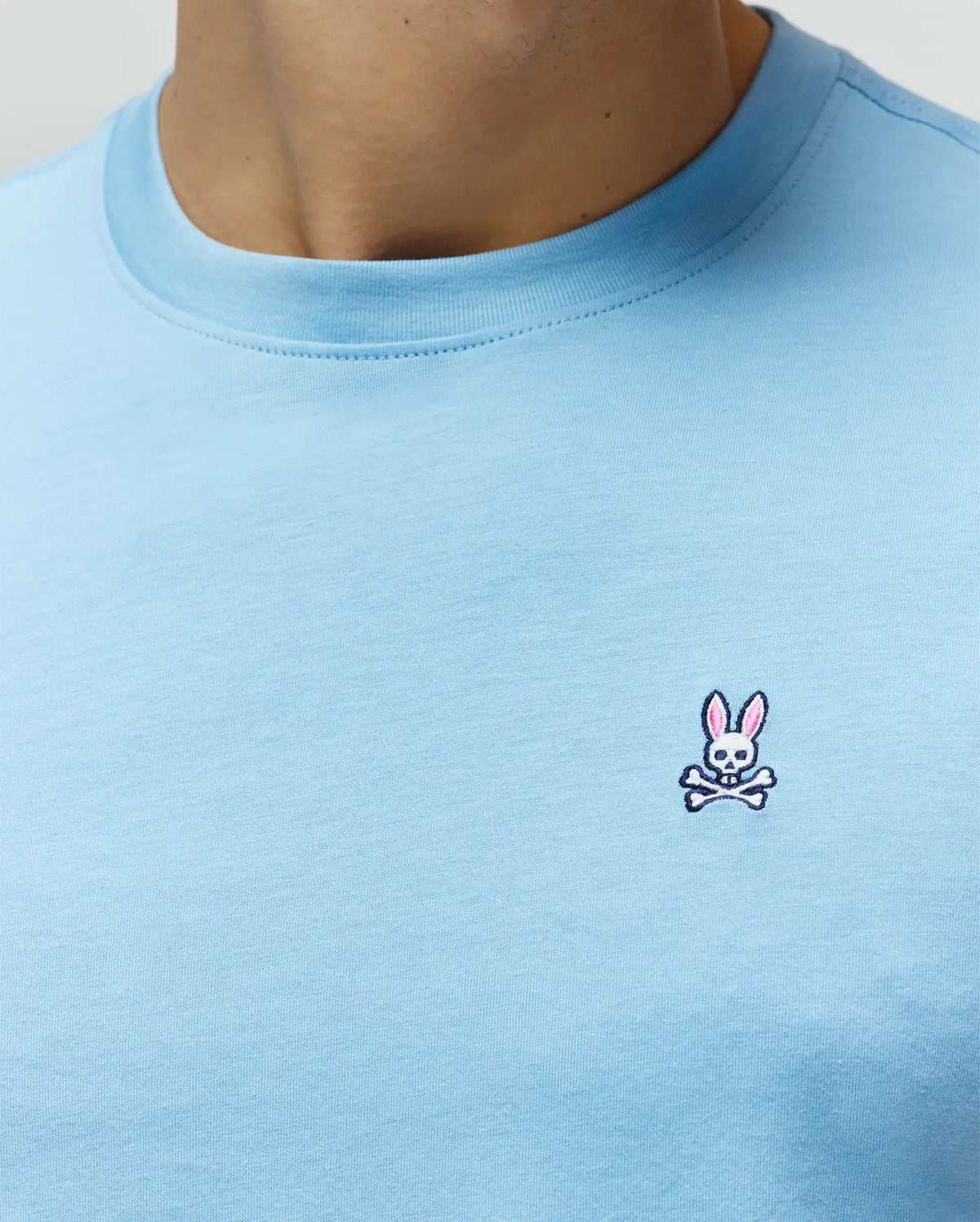 Mens Classic Crew Neck Tee in light blue jersey knit, featuring a small embroidered Psycho Bunny skull logo with pink ears on the left chest, classic fit.
