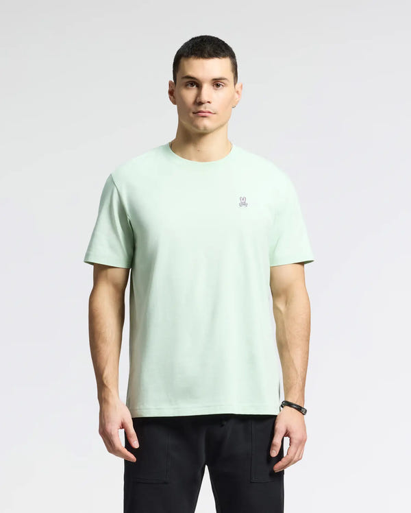 Against a plain backdrop, a person with short hair and a neutral expression wears Psycho Bunny's MENS CLASSIC CREW NECK TEE - B6U014F200 in light green Pima cotton, featuring an embroidered Bunny logo on the chest, paired with black pants.