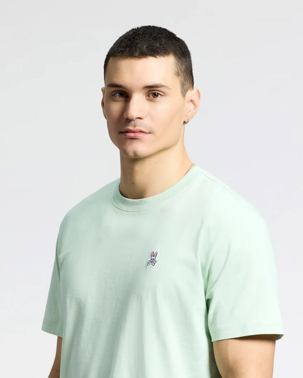 A person with short hair wears the Psycho Bunny MENS CLASSIC CREW NECK TEE (B6U014F200) in light green Pima cotton, featuring a small embroidered Bunny logo, standing against a gray background.