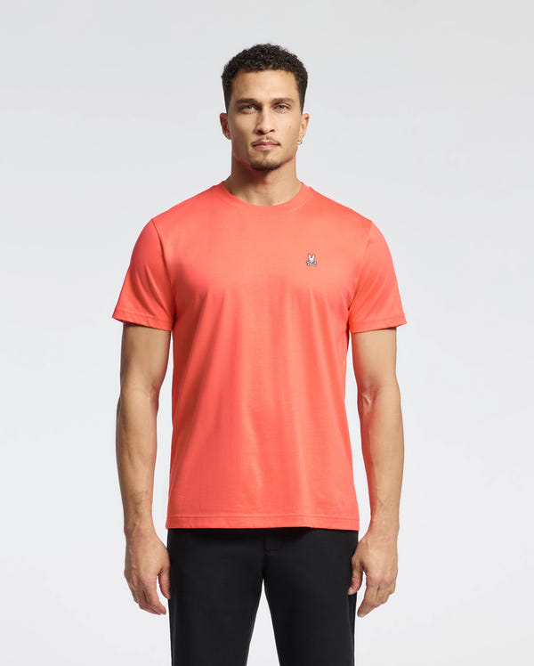 A person wearing a Psycho Bunny MENS CLASSIC CREW NECK TEE - B6U014F200 in bright orange stands against a plain white background, with short curly hair and looking directly at the camera.