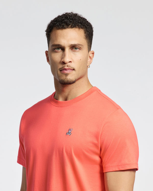 A man with short curly hair and facial hair wears the coral-colored Psycho Bunny MENS CLASSIC CREW NECK TEE - B6U014F200, featuring an embroidered bunny logo. He stands against a plain white background, exuding confidence as he looks at the camera.