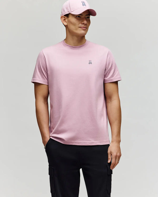 A person wears a Psycho Bunny MENS CLASSIC CREW NECK TEE - B6U014F200 in light pink with a matching cap, standing against a plain background and one hand in the pocket of black cargo pants.