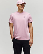 A person wears a Psycho Bunny MENS CLASSIC CREW NECK TEE - B6U014F200 in light pink with a matching cap, standing against a plain background and one hand in the pocket of black cargo pants.