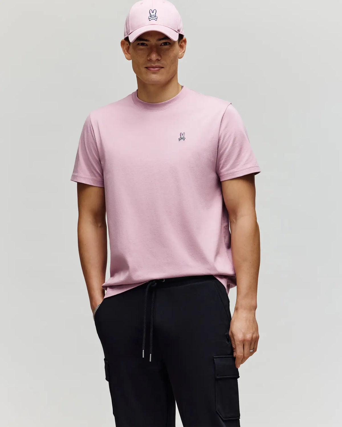 A man in a pink Psycho Bunny MENS CLASSIC CREW NECK TEE (B6U014F200) featuring a small embroidered bunny logo and black cargo pants stands against a plain background, wearing a matching pink cap with a neutral expression.