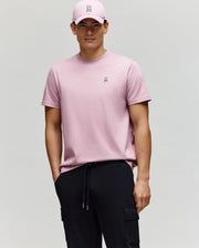 A man in a pink Psycho Bunny MENS CLASSIC CREW NECK TEE (B6U014F200) featuring a small embroidered bunny logo and black cargo pants stands against a plain background, wearing a matching pink cap with a neutral expression.