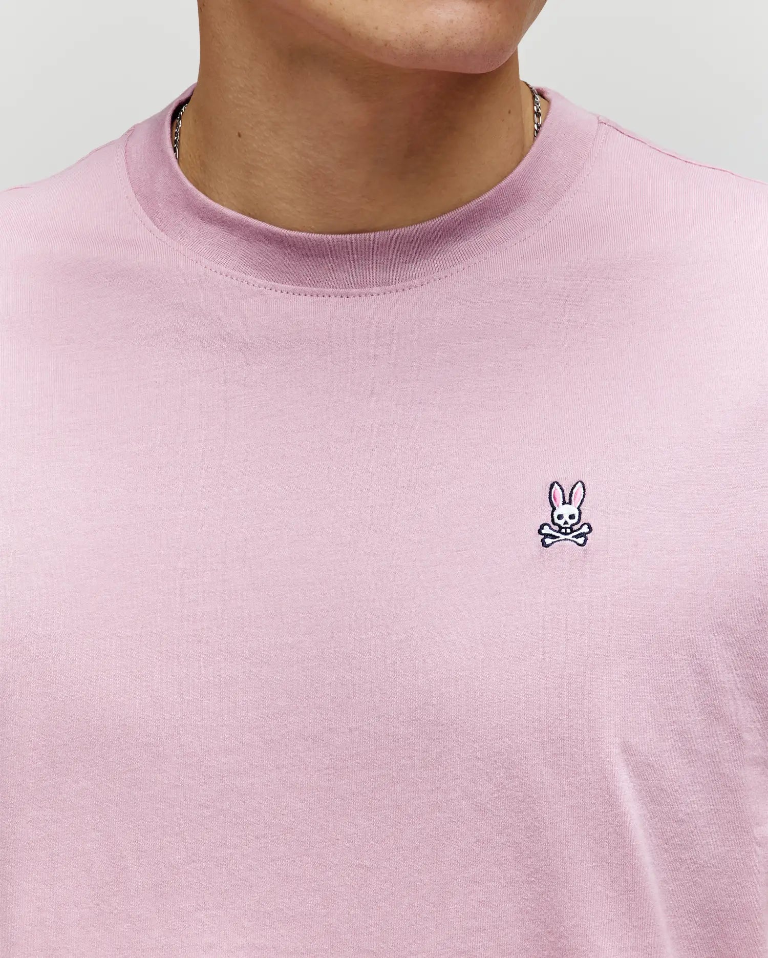 A person wears the Psycho Bunny MENS CLASSIC CREW NECK TEE - B6U014F200 in light purple, featuring a small embroidered bunny skull and crossbones logo on the chest. Face is partially out of frame.