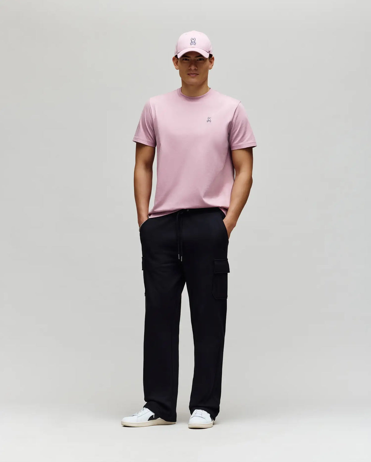 A man wears a light pink Psycho Bunny MENS CLASSIC CREW NECK TEE (B6U014F200), matching pink cap, black cargo pants, and white sneakers. He stands against a plain background, hands in pockets, looking straight ahead.