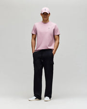 A man wears a light pink Psycho Bunny MENS CLASSIC CREW NECK TEE (B6U014F200), matching pink cap, black cargo pants, and white sneakers. He stands against a plain background, hands in pockets, looking straight ahead.