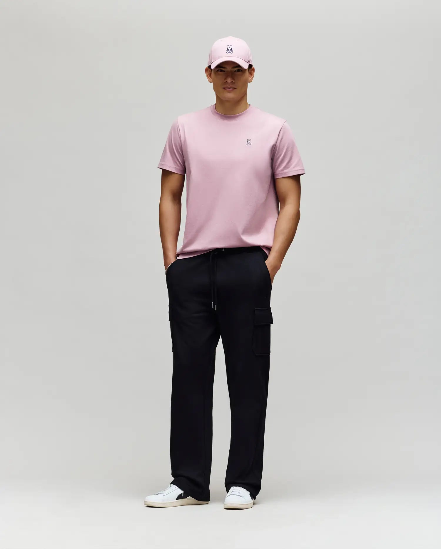 A man wears a light pink Psycho Bunny MENS CLASSIC CREW NECK TEE (B6U014F200), matching pink cap, black cargo pants, and white sneakers. He stands against a plain background, hands in pockets, looking straight ahead.