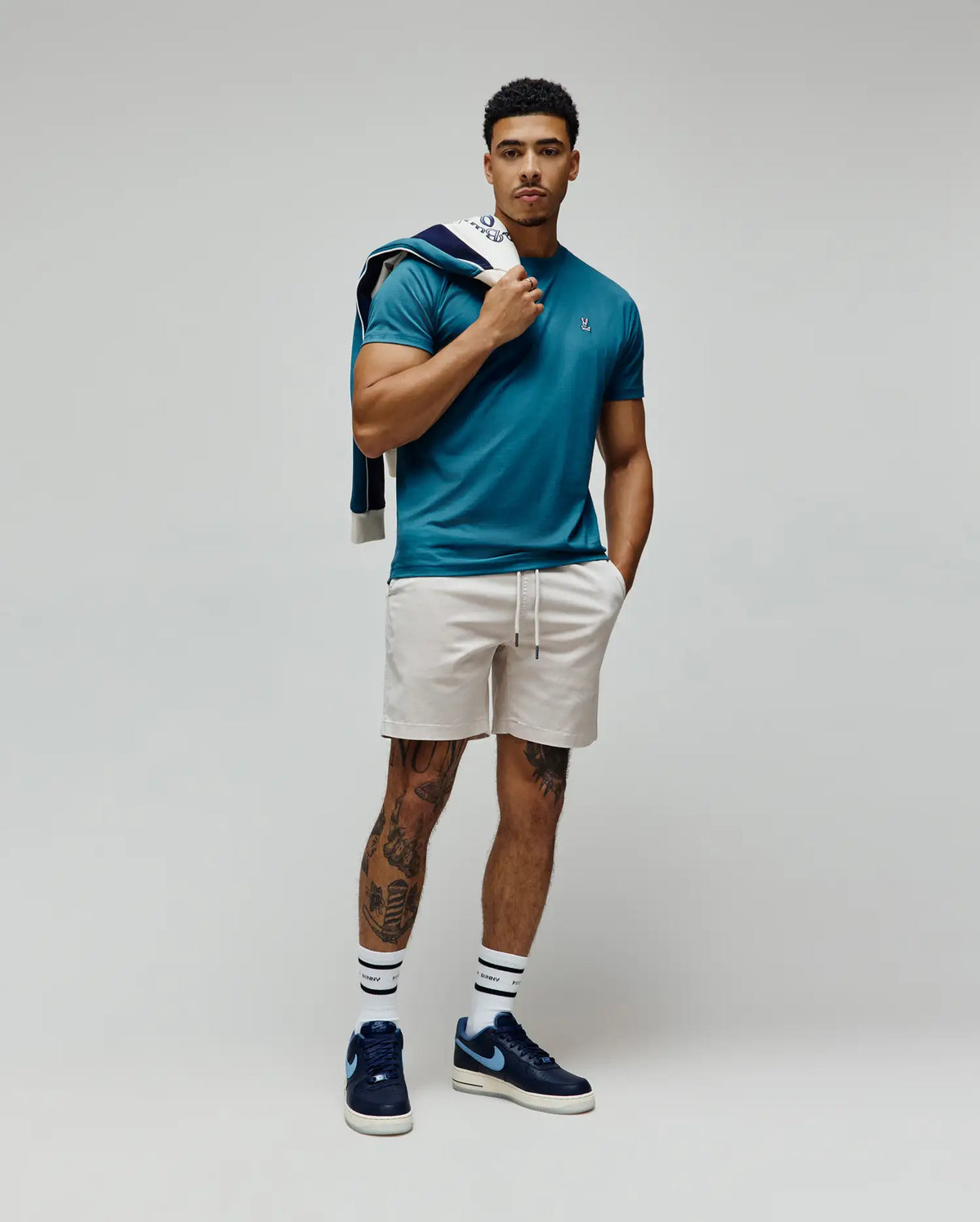 Mens Classic Crew Neck Tee in teal jersey with a slim fit and small embroidered Psycho Bunny skull logo on the left chest, paired with light beige shorts and a navy patterned jacket.