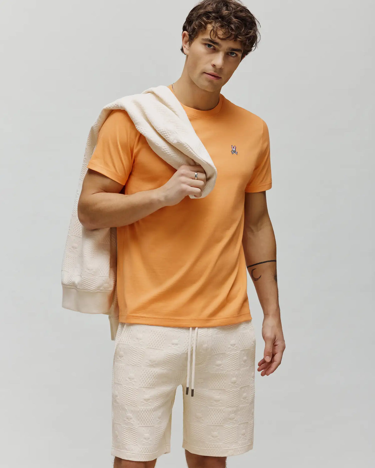 Vibrant orange MENS CLASSIC CREW NECK TEE with a small embroidered Psycho Bunny skull logo on the left chest, paired with cream textured knit shorts and a matching sweatshirt featuring an embossed skull pattern.