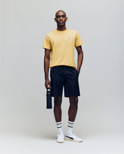 A person poses against a plain backdrop wearing the Psycho Bunny MENS CLASSIC CREW NECK TEE - B6U014F200 in vibrant yellow, paired with black shorts, striped white socks, white sneakers, and holding a black water bottle.