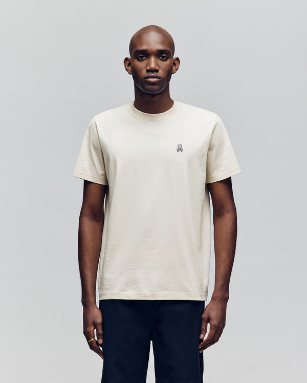 A man faces forward against a plain light gray background, wearing Psycho Bunny's MENS CLASSIC CREW NECK TEE (B6U014F200), an off-white pima cotton short-sleeve crewneck with a small embroidered logo on the left chest and black pants.
