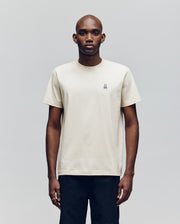 A man faces forward against a plain light gray background, wearing Psycho Bunny's MENS CLASSIC CREW NECK TEE (B6U014F200), an off-white pima cotton short-sleeve crewneck with a small embroidered logo on the left chest and black pants.