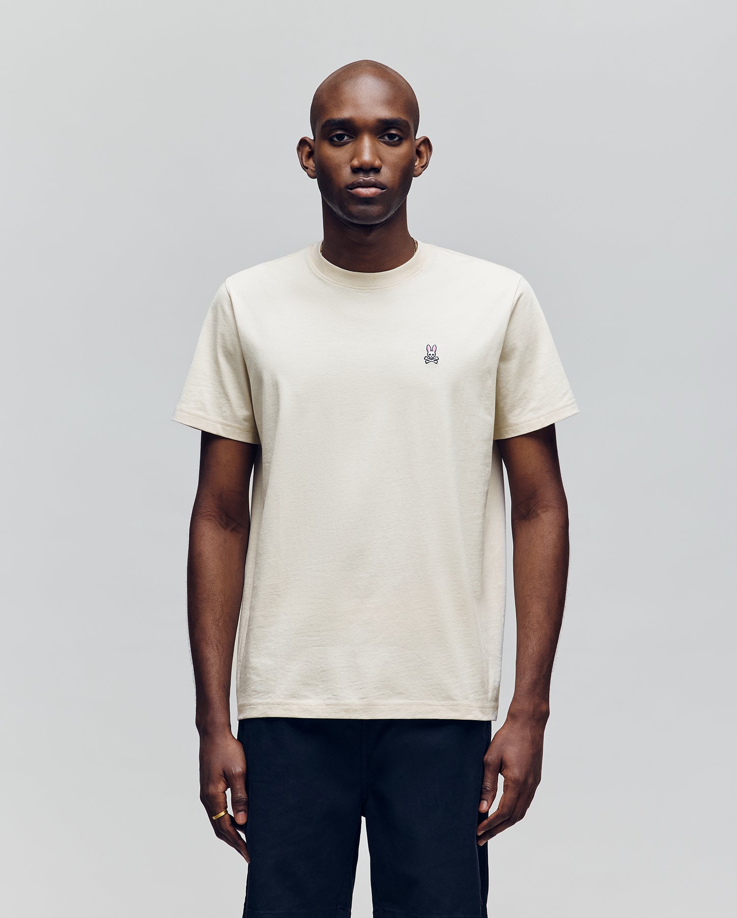 A man faces forward against a plain light gray background, wearing Psycho Bunny's MENS CLASSIC CREW NECK TEE (B6U014F200), an off-white pima cotton short-sleeve crewneck with a small embroidered logo on the left chest and black pants.