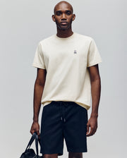 A man faces forward, wearing a cream Psycho Bunny MENS CLASSIC CREW NECK TEE (B6U014F200) with a small embroidered logo and black drawstring shorts. He holds a black bag in his left hand against a plain light gray background.