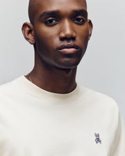 A man with a shaved head wears the Psycho Bunny MENS CLASSIC CREW NECK TEE (B6U014F200) in cream pima cotton, featuring a small pink rabbit with crossbones embroidered on the chest, standing against a neutral background.