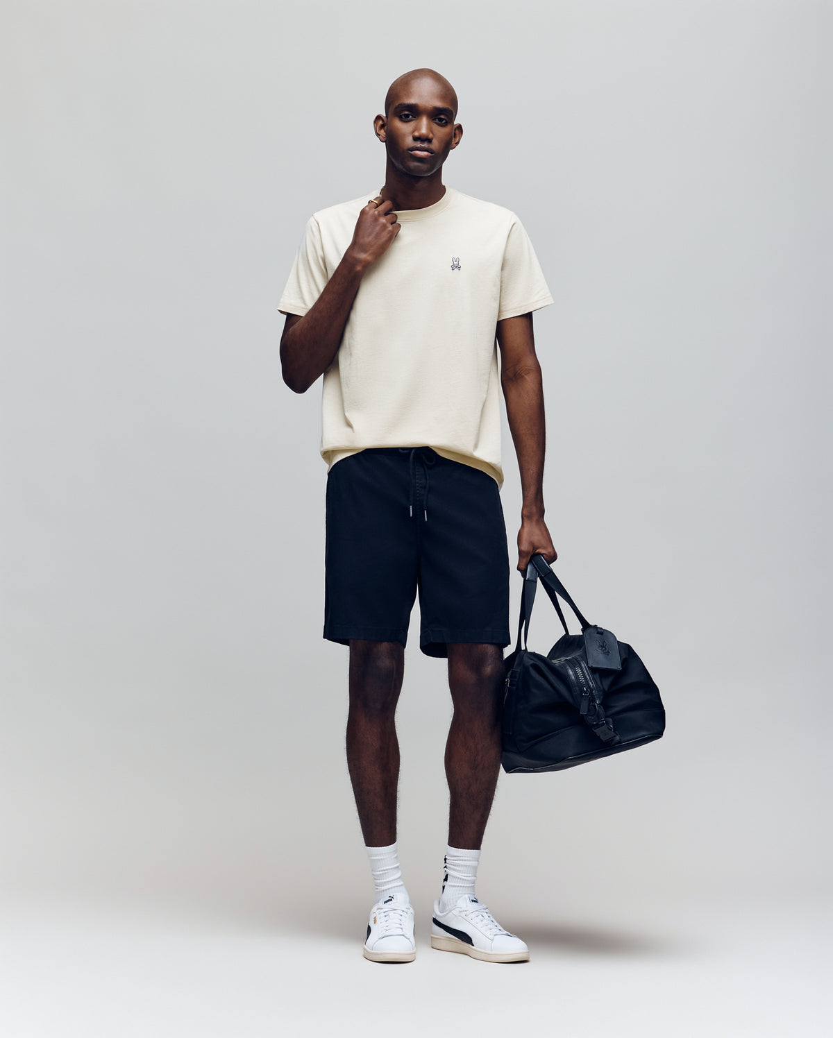 A man wearing the Psycho Bunny MENS CLASSIC CREW NECK TEE - B6U014F200 in beige, black shorts, white socks, and white sneakers stands holding a black duffel bag against a plain light grey background.