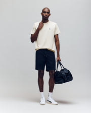A man wearing the Psycho Bunny MENS CLASSIC CREW NECK TEE - B6U014F200 in beige, black shorts, white socks, and white sneakers stands holding a black duffel bag against a plain light grey background.