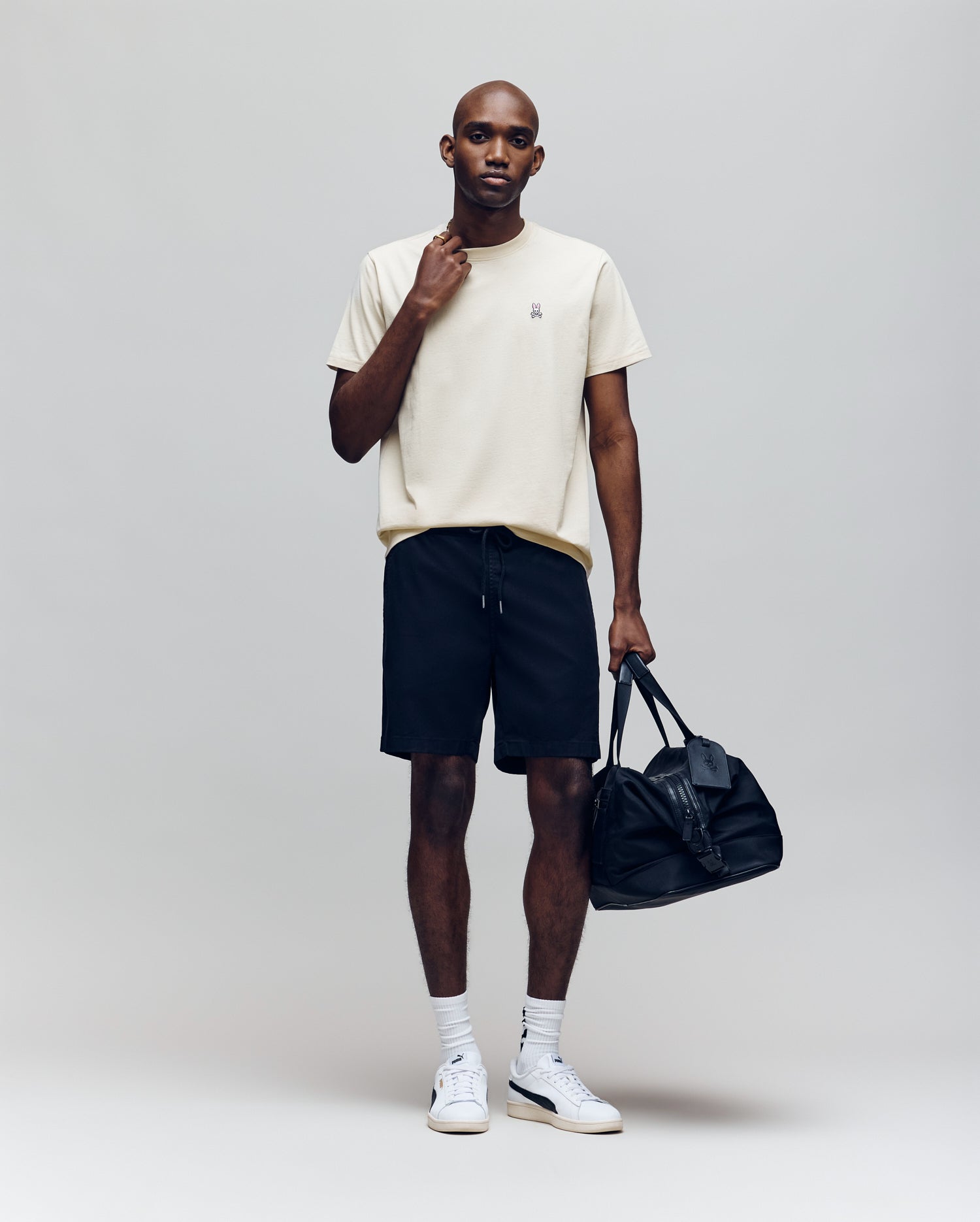 A man wearing the Psycho Bunny MENS CLASSIC CREW NECK TEE - B6U014F200 in beige, black shorts, white socks, and white sneakers stands holding a black duffel bag against a plain light grey background.