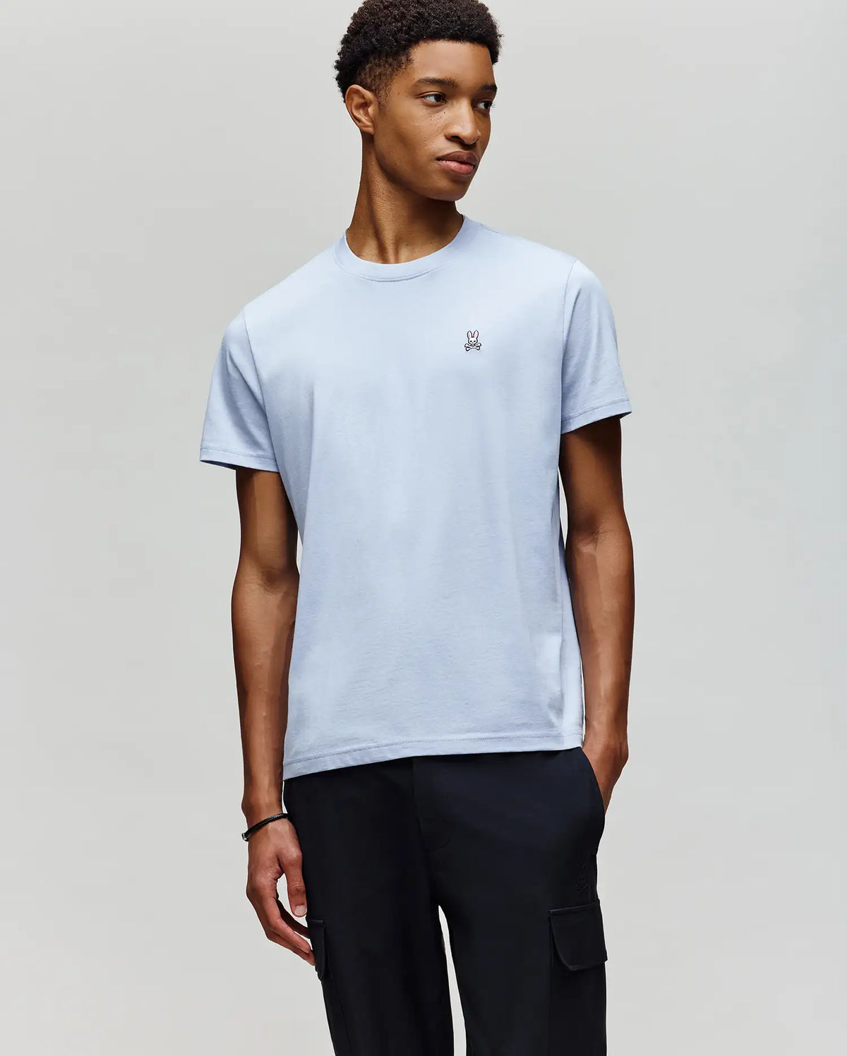 A young man stands against a gray background, wearing Psycho Bunny's MENS CLASSIC CREW NECK TEE (B6U014F200) in light blue Pima cotton with a small chest logo, paired with black cargo pants.
