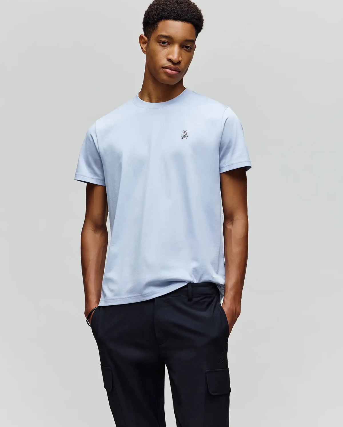 A young man stands with his hands in his pockets, wearing a Psycho Bunny MENS CLASSIC CREW NECK TEE - B6U014F200 in light blue and black pants, set against a plain light gray background.