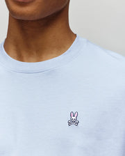 A person wearing the Psycho Bunny MENS CLASSIC CREW NECK TEE - B6U014F200 in premium light blue, featuring a small embroidered pink bunny and crossbones logo on the chest.
