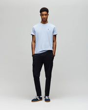 A young man stands against a light gray background, wearing the Psycho Bunny MENS CLASSIC CREW NECK TEE (B6U014F200) in light blue, paired with black cargo pants and navy sneakers. His hands are in his pockets and he looks straight ahead neutrally.