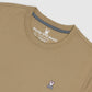 Close-up of a beige men's classic crew neck tee with the Psycho Bunny embroidered logo featuring a stylized rabbit head, positioned on the left chest area of the shirt.