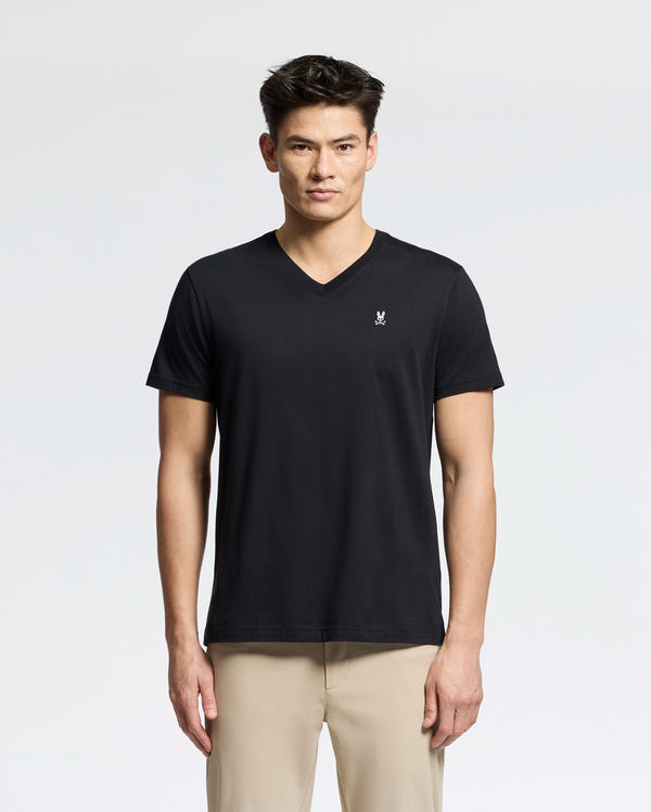 A person in the Psycho Bunny MENS CLASSIC V NECK TEE - B6U100ARPC, made of soft Pima cotton and featuring a small white chest logo, pairs it with light beige pants against a plain white background. They have a neutral expression while wearing this black tee.