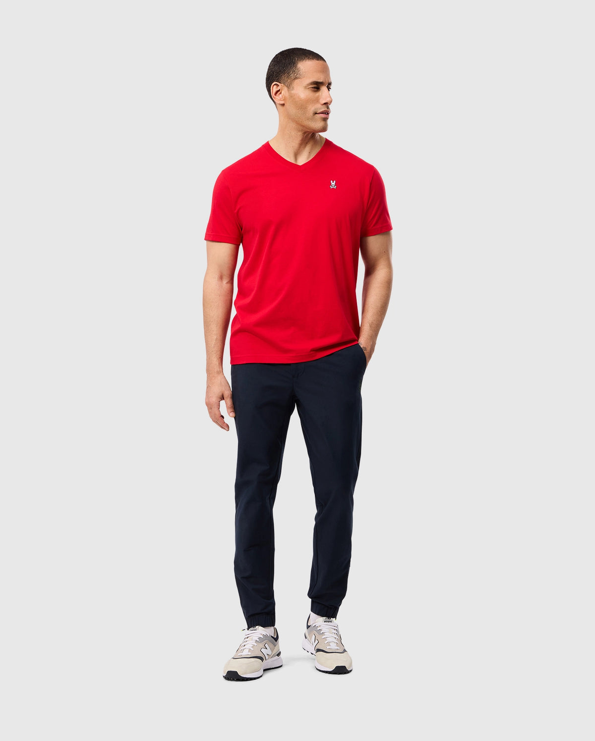 A person wearing a red Psycho Bunny MENS CLASSIC V NECK TEE (B6U100CRPC), paired with navy pants and white sneakers, stands against a plain background. They have short dark hair, are gazing to the side, and have one hand in their pocket.