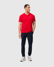 A person wearing a red Psycho Bunny MENS CLASSIC V NECK TEE (B6U100CRPC), paired with navy pants and white sneakers, stands against a plain background. They have short dark hair, are gazing to the side, and have one hand in their pocket.