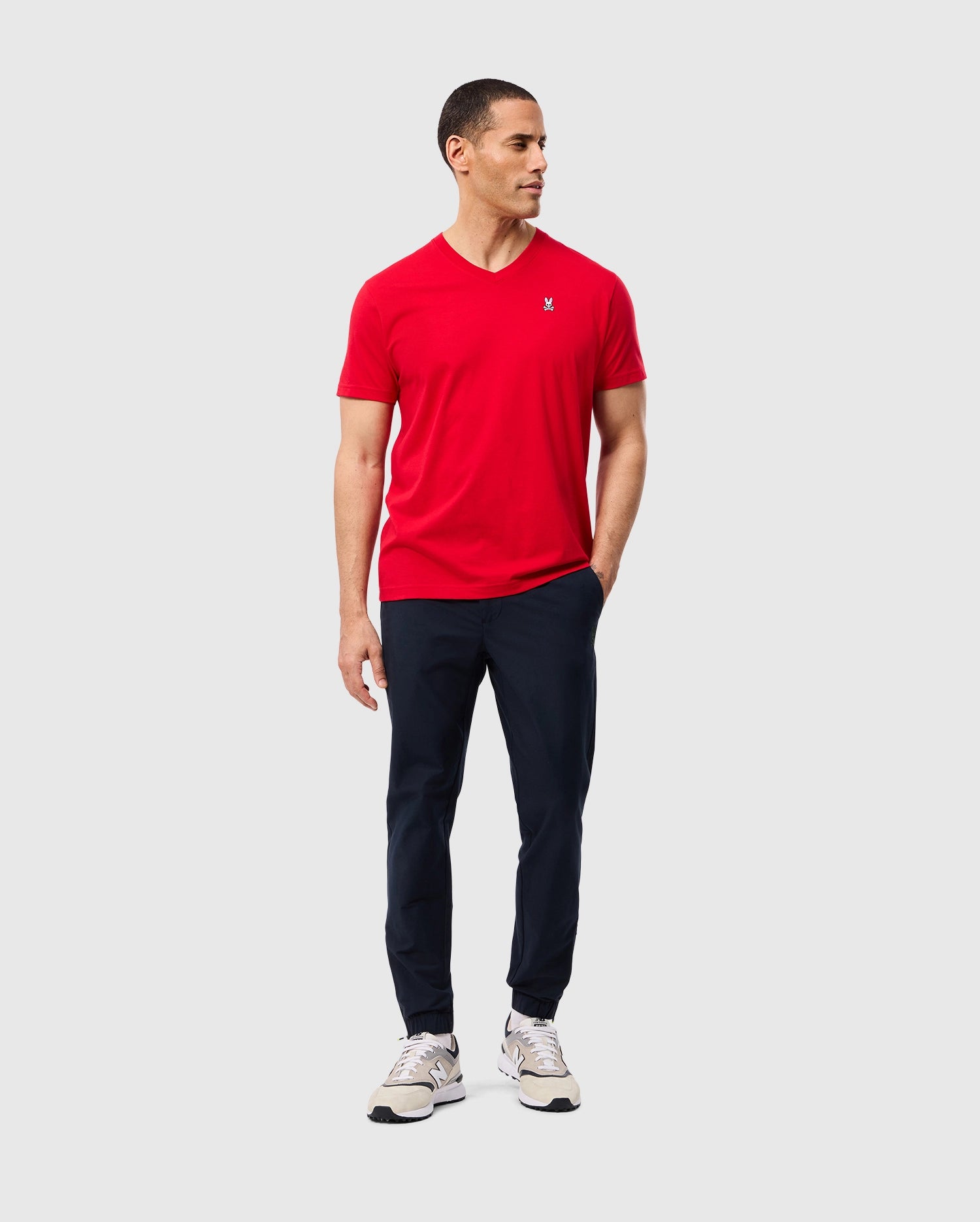 A person wearing a red Psycho Bunny MENS CLASSIC V NECK TEE (B6U100CRPC), paired with navy pants and white sneakers, stands against a plain background. They have short dark hair, are gazing to the side, and have one hand in their pocket.