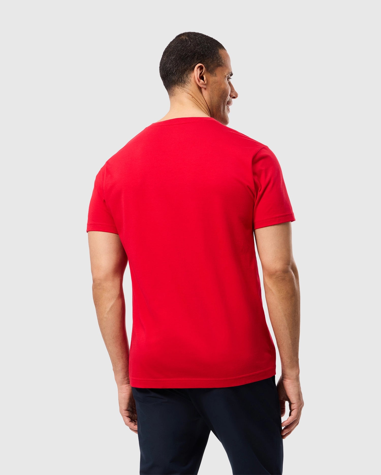A man with short hair is seen wearing the vibrant red MENS CLASSIC V-NECK TEE - B6U100CRPC, crafted from soft Pima cotton by Psycho Bunny, paired with dark pants. Positioned against a light gray backdrop and facing away from the camera, he subtly highlights the iconic Psycho Bunny logo on the back.