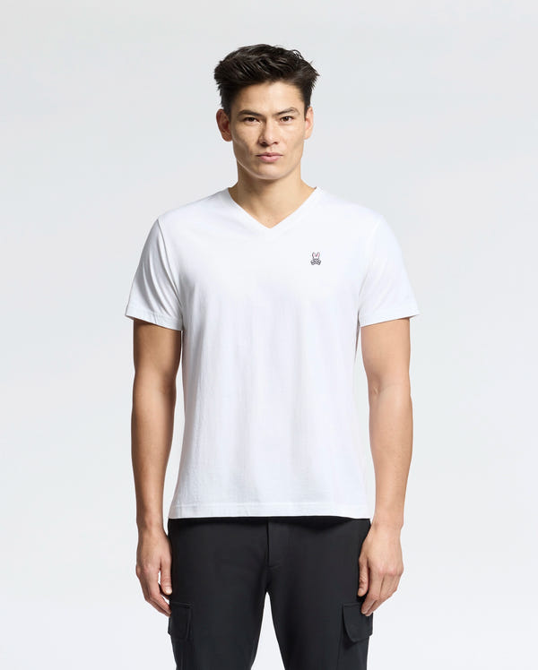 Against a light background, a person wears the Psycho Bunny Men's Classic V-Neck Tee in white with an embroidered logo, paired with black pants. The 100% Pima cotton tee complements their short dark hair as they look directly at the camera with a neutral expression.