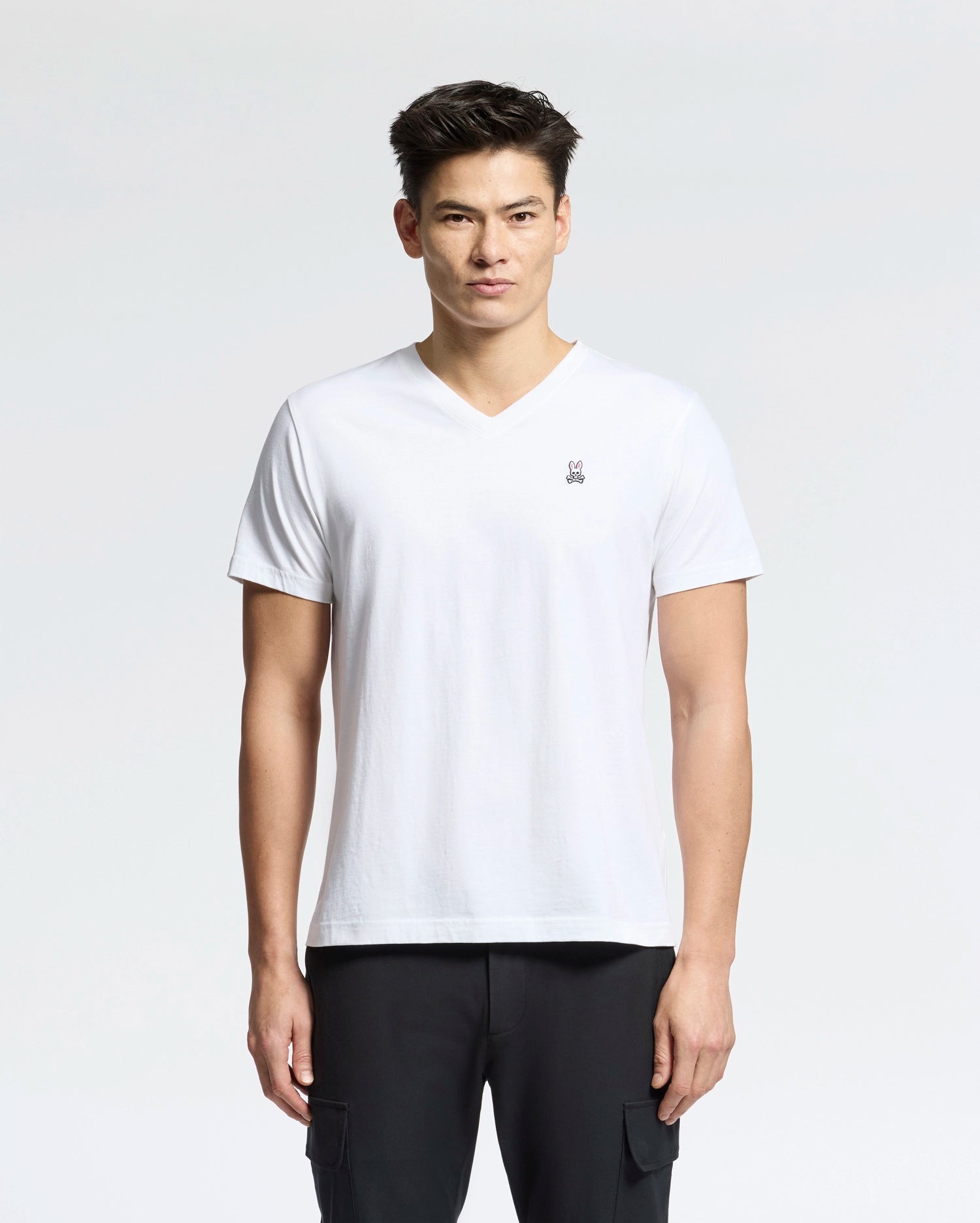 Against a light background, a person wears the Psycho Bunny Men's Classic V-Neck Tee in white with an embroidered logo, paired with black pants. The 100% Pima cotton tee complements their short dark hair as they look directly at the camera with a neutral expression.