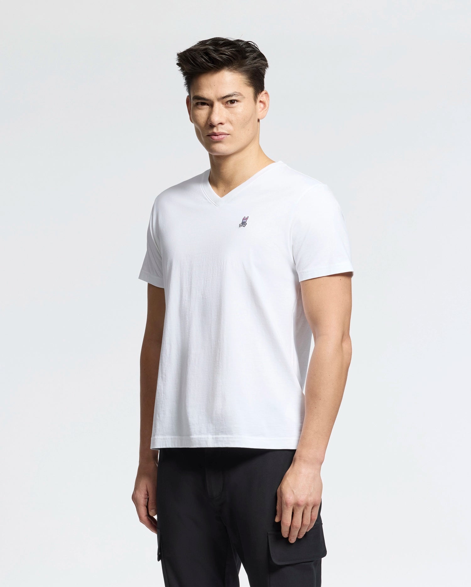 A person in Psycho Bunny's MENS CLASSIC V NECK TEE (B6U100ARPC), a white 100% Pima cotton tee with a small embroidered logo on the chest, stands against a plain backdrop in black pants.